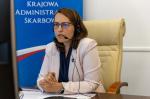 Przy biurku siedzi Szef Krajowej Administracji Skarbowej Magdalena Rzeczkowska i ma słuchawki na uszach oraz mikrofon