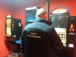 W pomieszczeniu stoją trzy automaty do gier hazardowych. Obok stoi mężczyzna w mundurze.