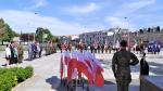 Uroczysty apel na placu przed Pomnikiem Czynu Legionowego w Kielcach. Na pierwszym planie biało-czerwone flagi narodowe oraz żołnierz w pozycji na baczność. W tle uczestnicy uroczystości oraz poczty sztandarowe różnych instytucji.